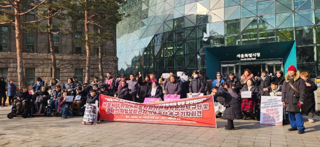 25년 2월 14일 진행된 기자회견 진행사진. 서울시청을 등지고 장애인권활동가들이 모여있다. 붉은 바탕의 현수막이 중앙에 있고 그 옆에 놓인 쓰레기봉투에는 '서울시 장애인활동지원기관 재지정심사' 문구가 부착되어있다.