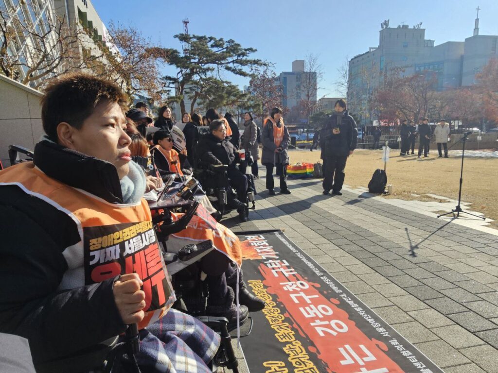 24년 12월 23일 진행 기자회견 의 진행사진. 강동구청을 등지고 휠체어 이용 활동가들이 일렬로 있다. 장애여성활동가가 굳은 얼굴로 정면을 보고 있다.