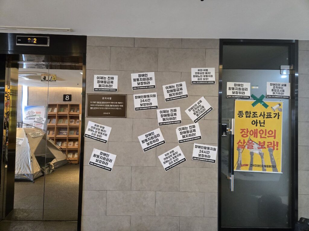 <사진 2. 10월 16일 강동하남지사 점거 농성 사진. 국민연금공단 강동하남지사 엘리베이터에 ‘종합조사표가 아닌 장애인의 삶을 보라!, 이제는 진짜 장애등급제 폐지하라!’ 등의 피켓이 가득 붙어있다.>