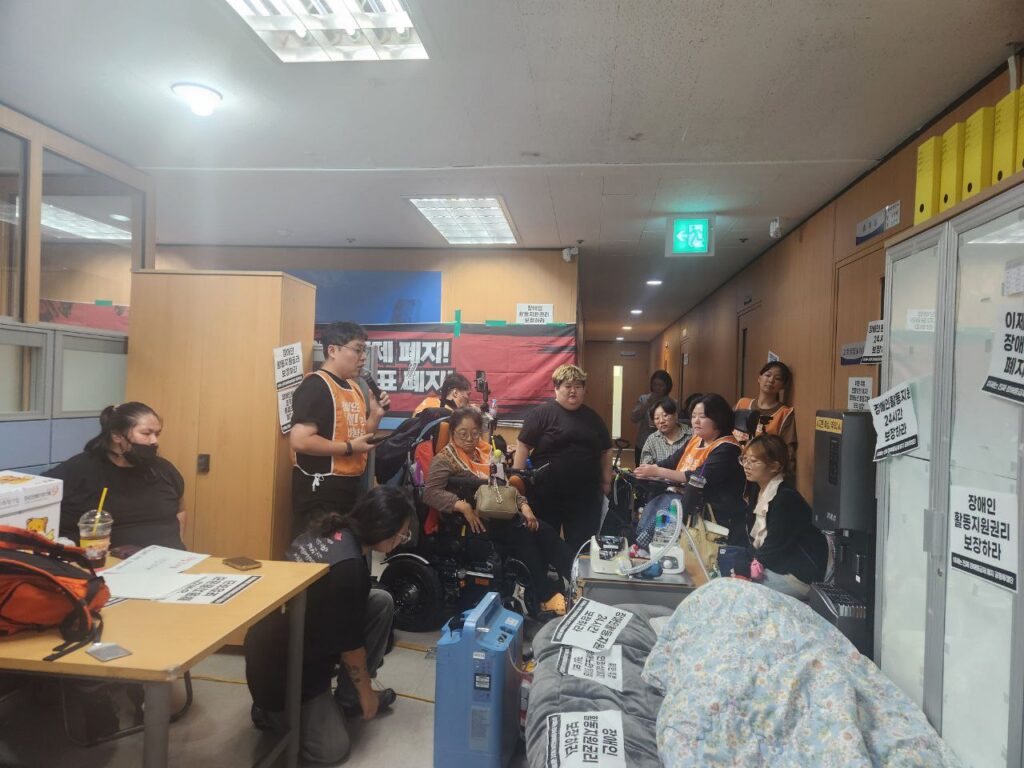 <사진 1. 10월 16일 강동하남지사 점거 농성 사진. 국민연금공단 강동하남지사 사무실에서 전장연 소속 활동가들이 침대에 누워있는 A님을 바라보고 있다. 한 활동가가 투쟁 상황을 동지들에게 공유하고 있다.> 