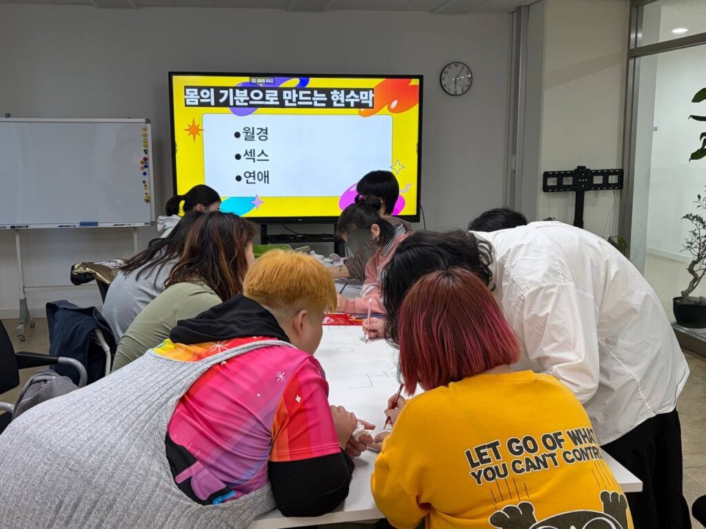 (사진 1) 모임 첫날, 함께 책상에 둘러앉아 전지에 그림을 그리고 있다. TV 화면에 “몸의 기분으로 만드는 현수막, 월경, 섹스, 연애”라고 적혀 있다.