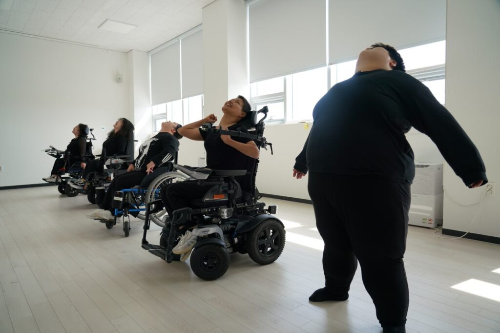 사진 1. 제주 몸이동(異動) 워크숍 첫째날. 장애여성들이 휠체어에 앉거나 바닥에 서서 상체를 뒤로 젖히거나 고개를 들며 몸을 풀고 있다.